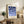 May include: Framed geometric art print with blue circular and semicircular shapes arranged in a grid pattern on cream background, displayed on rustic wooden sideboard. Room setting shows brown leather chair, small potted fern, ceramic vase, and stacked books against white wall with natural lighting.