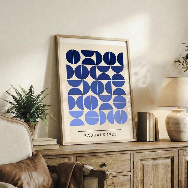 May include: Framed geometric art print with blue circular and semicircular shapes arranged in a grid pattern on cream background, displayed on rustic wooden sideboard. Room setting shows brown leather chair, small potted fern, ceramic vase, and stacked books against white wall with natural lighting.