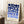 May include: Framed geometric art print with blue circles and semicircles in gradient shades from navy to light blue on cream background, text reading "BAUHAUS 1923" at bottom. Wooden frame leaning against white wall corner next to small wooden table with white coffee cup and saucer, cream knit throw blanket visible on white textured rug.