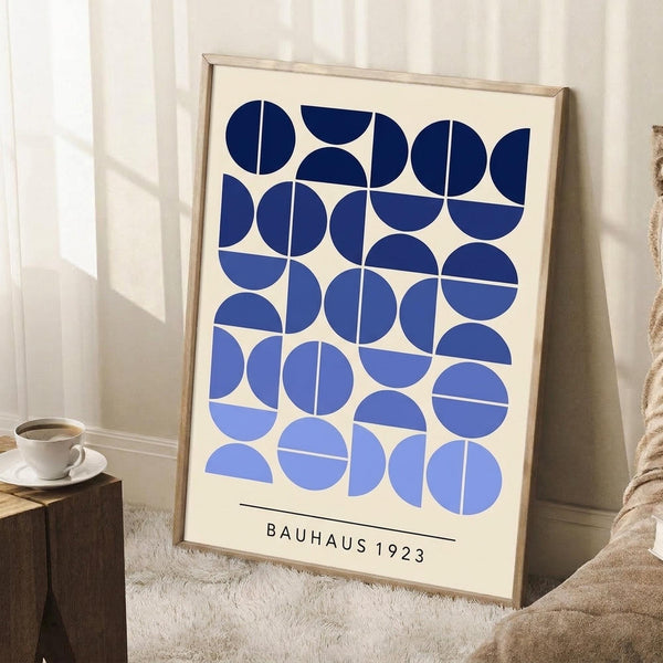 May include: Framed geometric art print with blue circles and semicircles in gradient shades from navy to light blue on cream background, text reading "BAUHAUS 1923" at bottom. Wooden frame leaning against white wall corner next to small wooden table with white coffee cup and saucer, cream knit throw blanket visible on white textured rug.