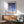 May include: Framed geometric art print with blue circles and semicircles in varying shades from navy to light blue on cream background, displayed above mid-century modern wooden credenza with white ceramic lamp, next to sage green upholstered armchair with wooden frame in sunlit room with white walls and wooden floors.