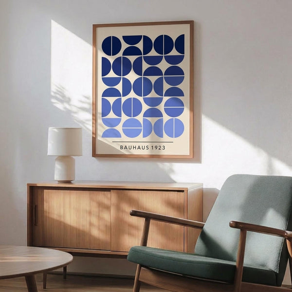 May include: Framed geometric art print with blue circles and semicircles in varying shades from navy to light blue on cream background, displayed above mid-century modern wooden credenza with white ceramic lamp, next to sage green upholstered armchair with wooden frame in sunlit room with white walls and wooden floors.