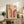 May include: Framed Bauhaus poster with geometric rainbow arcs in orange and burgundy tones on beige background, displayed on rustic wood sideboard next to fern plant, leather chair, ceramic vase, and stacked books in warm neutral living room setting