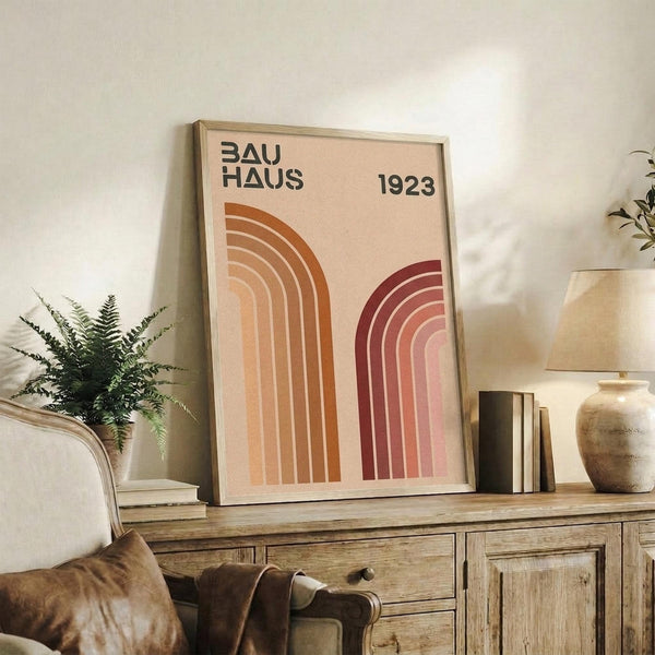 May include: Framed Bauhaus poster with geometric rainbow arcs in orange and burgundy tones on beige background, displayed on rustic wood sideboard next to fern plant, leather chair, ceramic vase, and stacked books in warm neutral living room setting