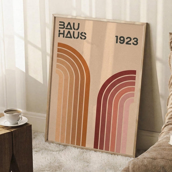 May include: Framed Bauhaus poster with geometric rainbow arcs in warm orange and burgundy tones on beige background, displaying "BAUHAUS 1923" text. Poster leans against white wall corner next to wooden side table with white coffee cup, cream textured rug, and beige knit throw blanket creating cozy modern interior styling.