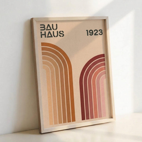 May include: Framed vintage-style poster with beige background featuring "BAUHAUS 1923" text and two curved rainbow arches in warm orange and burgundy tones. The minimalist geometric design sits in a light wood frame, leaning against a white wall on what appears to be a light surface.