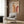 May include: Beige poster with "BAUHAUS 1923" text and geometric rainbow arches in orange and burgundy tones, displayed on wood paneled wall above cream upholstered chair with geometric patterned pillow, brass floor lamp and small round table with open book nearby