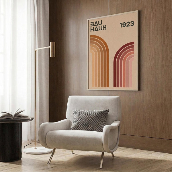 May include: Beige poster with "BAUHAUS 1923" text and geometric rainbow arches in orange and burgundy tones, displayed on wood paneled wall above cream upholstered chair with geometric patterned pillow, brass floor lamp and small round table with open book nearby