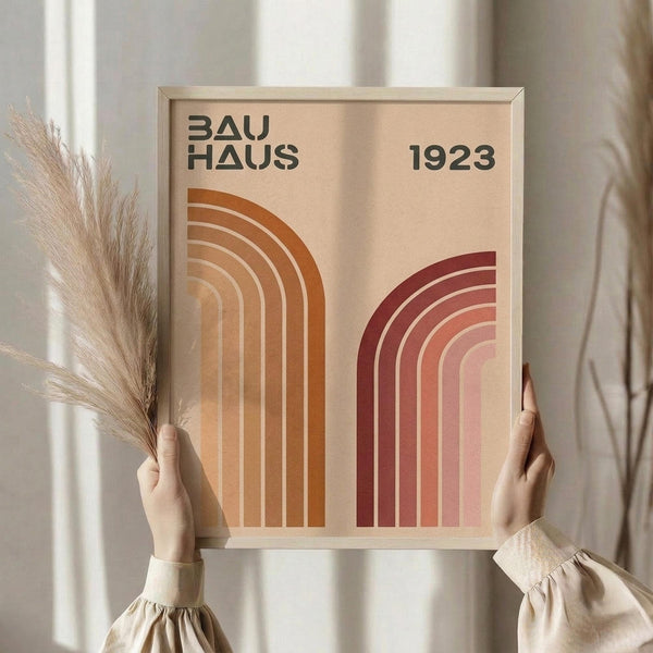 May include: Person in beige clothing holding framed Bauhaus poster featuring two geometric rainbow arches in orange and burgundy tones on cream background, displayed against neutral wall with dried pampas grass stems visible to the left side.
