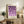 May include: Framed geometric poster with purple circles and semicircles in a grid pattern on cream background, displayed on rustic wooden sideboard next to fern plant, ceramic vases, and books in neutral living room setting