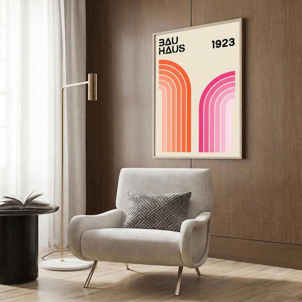 May include: Modern living room scene showing a framed geometric poster with orange and pink rainbow arches on cream background, text reading "BAUHAUS 1923" in black letters. Light wood frame hangs on dark wood paneled wall above beige upholstered armchair with geometric patterned pillow. Gold floor lamp and black side table with open book complete the mid-century modern interior styling.
