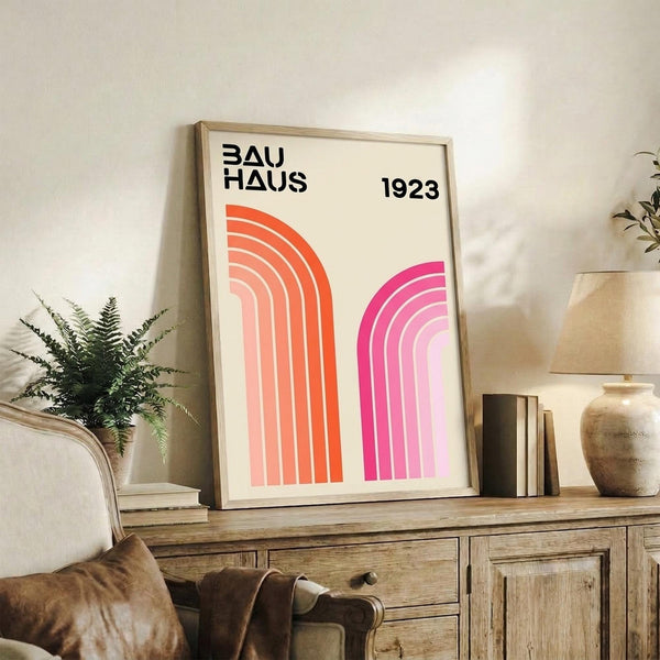 May include: Framed Bauhaus poster with geometric rainbow arcs in orange and pink gradients, displayed on rustic wood sideboard with books, ceramic vases, and fern plant in neutral-toned living room setting with beige chair and textured throw pillows