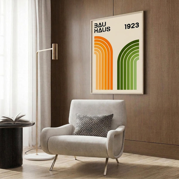 May include: Modern living room with beige armchair and geometric patterned pillow, gold floor lamp, dark round side table with open book, and framed poster on wood-paneled wall. Poster features orange and green curved arches with "BAUHAUS 1923" text on cream background in light wooden frame.