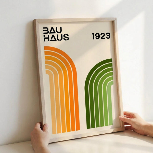 May include: Two hands holding a framed poster with orange and green geometric rainbow arches on cream background. The poster displays "BAUHAUS 1923" in black text at the top. The wooden frame has a light natural finish, and the image is photographed against a white wall with soft lighting creating subtle shadows.