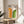 May include: Framed poster with cream background featuring orange and green geometric arches made of concentric curved lines, displayed on light wood round side table next to beige upholstered chair. Natural sunlight creates soft shadows on white wall behind. Green plant leaves visible in corner. Modern minimalist interior styling with neutral tones.