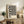 May include: Black and white abstract wavy stripes poster with "BAUHAUS" text in wooden frame, displayed on rustic wood sideboard with fern plant, ceramic vase, stacked books, and brown leather chair in neutral-toned living room setting