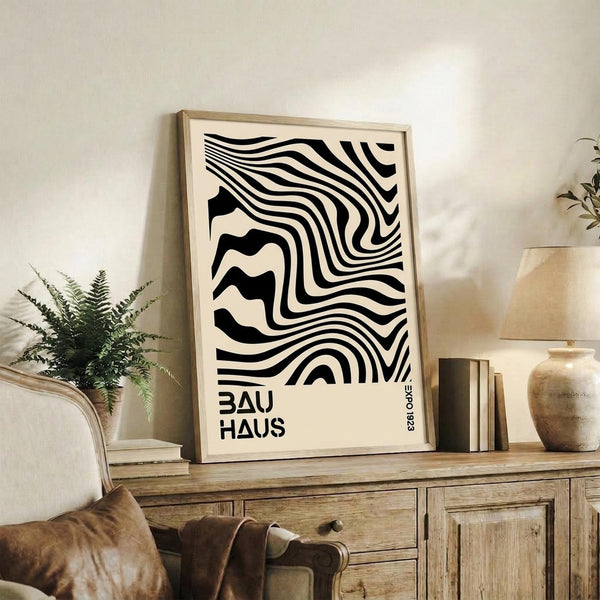 May include: Black and white abstract wavy stripes poster with "BAUHAUS" text in wooden frame, displayed on rustic wood sideboard with fern plant, ceramic vase, stacked books, and brown leather chair in neutral-toned living room setting