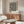 May include: Black and white wavy distorted stripes poster in wooden frame hanging above walnut credenza in modern living room. White sculptural chair and glass oval coffee table visible. Cream colored walls with natural lighting from window.