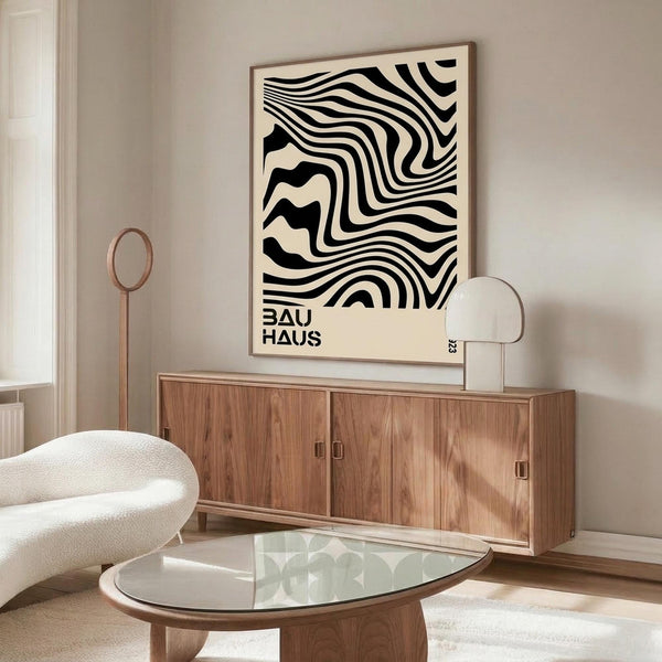 May include: Black and white wavy distorted stripes poster in wooden frame hanging above walnut credenza in modern living room. White sculptural chair and glass oval coffee table visible. Cream colored walls with natural lighting from window.
