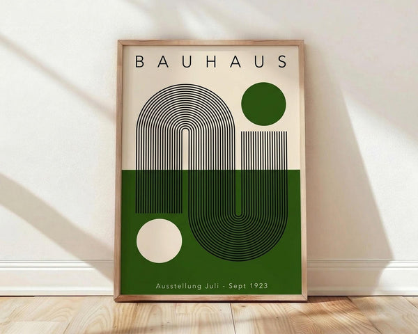 May include: Framed Bauhaus-style poster with geometric design featuring black concentric lines forming curved shapes, green and cream color blocks, small circles, and "Ausstellung Juli - Sept 1923" text, displayed on light wooden floor against white wall with natural lighting shadows