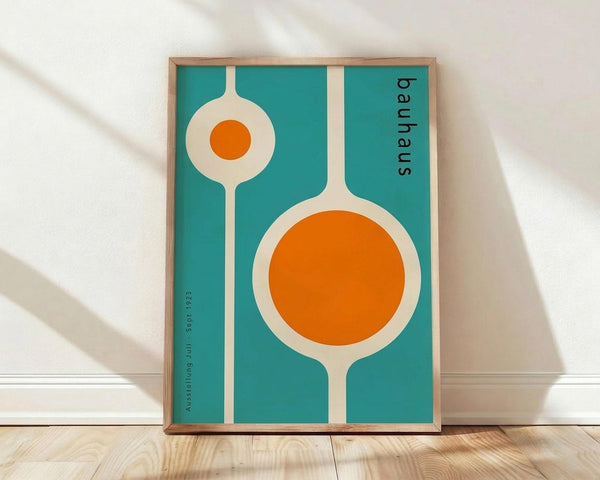 May include: Framed vintage-style Bauhaus exhibition poster with turquoise background featuring two cream-colored spoon or utensil shapes with bright orange circular centers, one large and one small. Black text reading "bauhaus" vertically along right edge and "Ausstellung Juli Sept 1923" along left side. Natural wood frame sitting on light wooden floor against white wall with soft shadows.