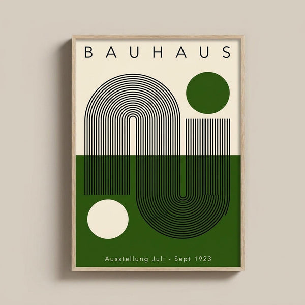 May include: Framed Bauhaus-style poster print hanging on white wall. Design features geometric black line patterns forming curved shapes, green and cream color blocks, two circles (one green, one cream), and "BAUHAUS" text at top with "Ausstellung Juli - Sept 1923" at bottom. Light wood frame surrounds the minimalist modernist artwork.