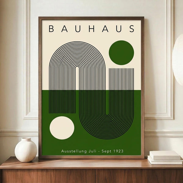 May include: Framed Bauhaus-style poster with geometric black line art forming curved shapes on cream and green color-blocked background, displayed on wooden sideboard next to white ceramic vase and stacked books in modern interior with white paneled walls.
