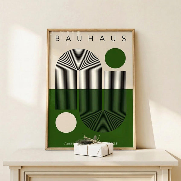 May include: Framed Bauhaus-style poster with geometric design featuring black concentric lines forming arch and circle shapes, green and cream color blocks, dark green circle, displayed on light wood surface next to small wrapped gift with twine and evergreen sprig against cream wall.