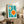 May include: Framed Bauhaus-style poster with teal background and white geometric shapes containing orange circles, displayed on rustic wooden sideboard alongside fern plant, ceramic vase, and stacked books in neutral-toned living room setting