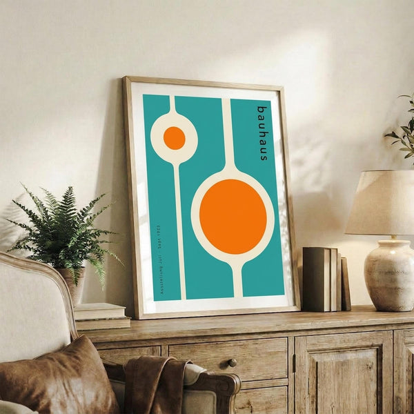 May include: Framed Bauhaus-style poster with teal background and white geometric shapes containing orange circles, displayed on rustic wooden sideboard alongside fern plant, ceramic vase, and stacked books in neutral-toned living room setting