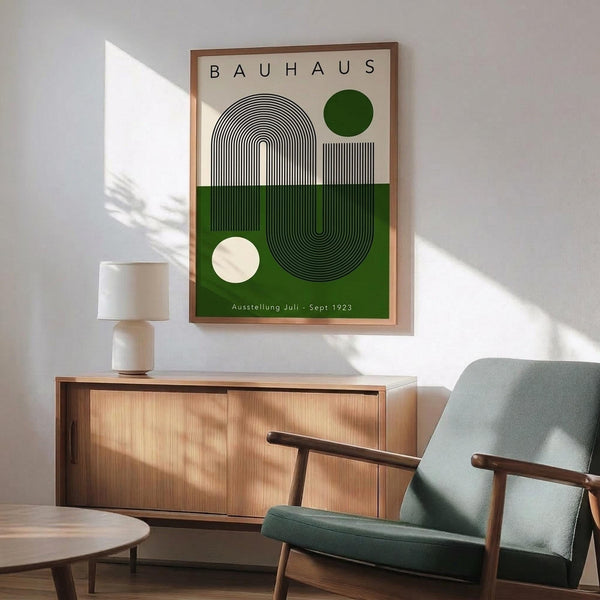 May include: Bauhaus-style poster print with geometric design featuring black and white concentric lines forming a U-shape, dark green and cream colored circles, and green lower section with "Ausstellung Juli - Sept 1923" text. Shown framed in thin wood frame above wooden sideboard with ceramic table lamp, positioned next to mid-century modern teal upholstered armchair with wooden frame. Natural sunlight casts shadows across white wall in minimalist interior setting.