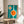 May include: Modernist poster with turquoise background featuring white geometric shapes - a large circle and smaller elongated oval, both filled with bright orange centers. Black "bauhaus" text appears vertically on the right side. The framed artwork sits on a round wooden side table next to a beige upholstered chair, with natural sunlight casting shadows and a plant visible in the corner of the minimalist room setting.