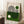 May include: Framed Bauhaus-style poster with geometric design featuring curved black lines forming abstract shapes, green and cream color blocks, white and green circles, displayed on white wooden floor against white wall. Coffee cup on wooden block visible nearby, with beige textured throw blanket in corner.