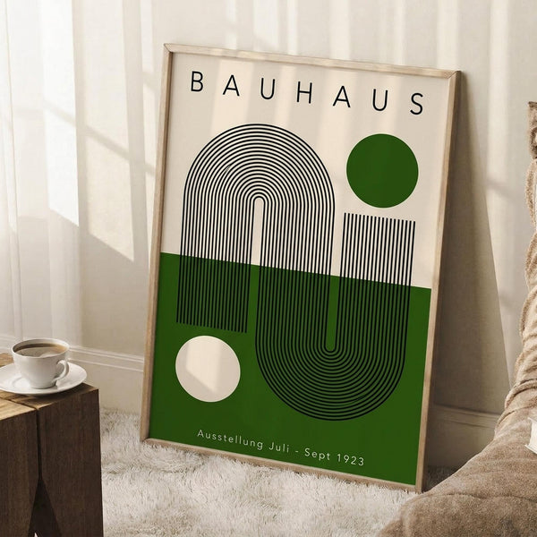 May include: Framed Bauhaus-style poster with geometric design featuring curved black lines forming abstract shapes, green and cream color blocks, white and green circles, displayed on white wooden floor against white wall. Coffee cup on wooden block visible nearby, with beige textured throw blanket in corner.