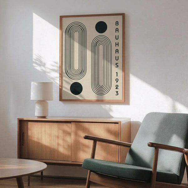 May include: Framed geometric Bauhaus poster with curved parallel lines forming abstract shapes and two black circles, displaying "BAUHAUS 1923" vertically. Shown in modern room setting with mid-century furniture including sage green upholstered chair with wooden arms, warm wood sideboard, white ceramic lamp, and round coffee table. Natural sunlight creates dramatic shadows across white walls.