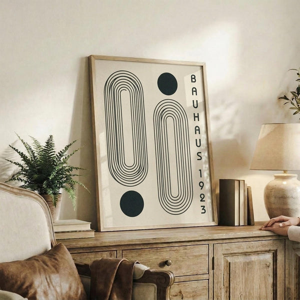 May include: Framed geometric art print with black line drawings of rounded rectangular shapes and circles, featuring "BAUHAUS 1923" text in vertical letters. Light wood frame displayed on rustic wooden sideboard alongside books, ceramic vase, and potted fern. Cream-colored wall background with beige armchair visible in cozy modern living room setting.
