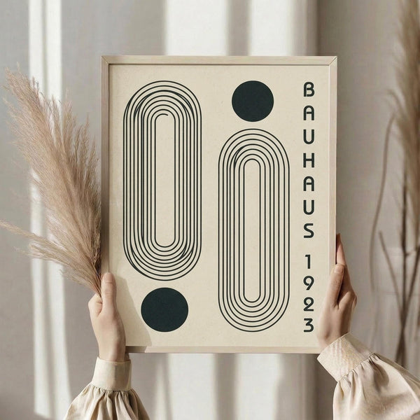 May include: Person holding framed geometric art print featuring two elongated oval shapes made of concentric black lines, two dark green circles, and vertical text reading "BAUHAUS 1923" on cream background. Light wood frame, neutral beige clothing, dried pampas grass visible, soft natural lighting against white curtained window.