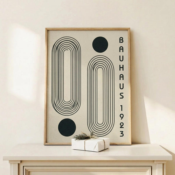 May include: Framed poster with geometric black line art featuring concentric oval and rectangular shapes, two solid black circles, and vertical text reading "BAUHAUS 1923" on cream background. Natural wood frame sits on light wooden surface next to small white wrapped gift with greenery. Clean minimalist room setting with white walls.
