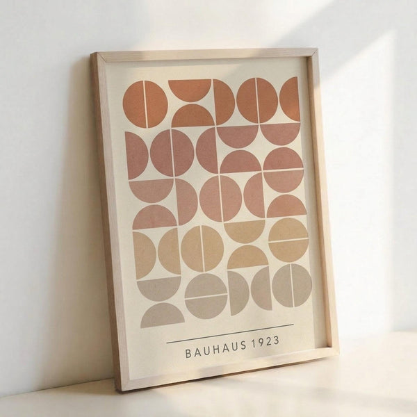 Framed geometric art print featuring circles and semicircles in warm terracotta, rust, peach, tan, and beige tones arranged in a grid pattern on cream background. Text reads "BAUHAUS 1923" at bottom. Light wood frame sits against white wall with soft shadows, showing the poster's earth-toned minimalist design inspired by Bauhaus aesthetic.