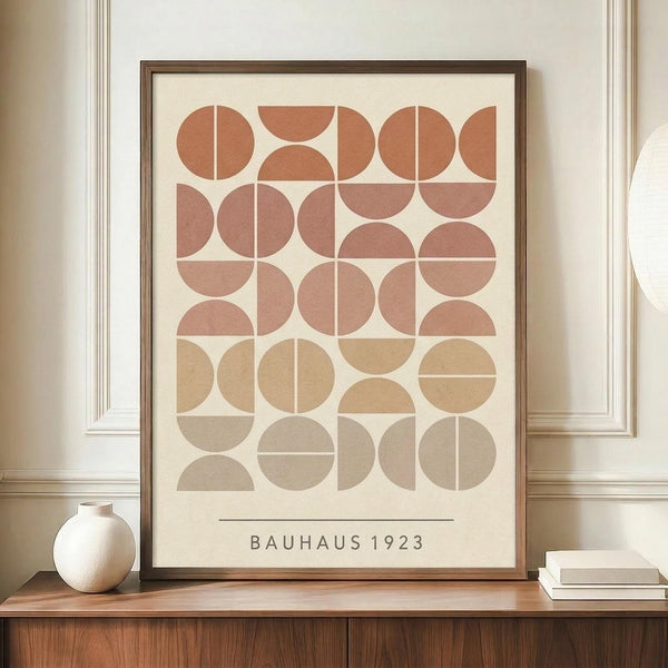 May include: Framed geometric poster with earth-toned circles and semicircles arranged in a grid pattern, featuring warm rust, terracotta, and beige colors on cream background. Text reads "BAUHAUS 1923" at bottom. Displayed on wooden sideboard with small white ceramic vase and stacked books in elegant room with white paneled walls.