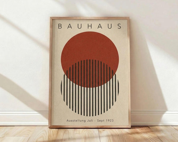 May include: A framed Bauhaus-style poster with geometric design featuring a rust-red circle overlapping a black and white striped circle on cream background, displayed in light wood frame against white wall on natural wood floor with soft shadow patterns.