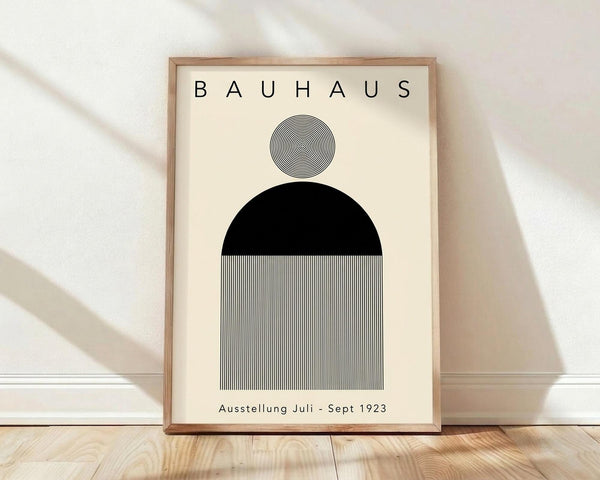 May include: Framed minimalist Bauhaus poster with geometric design featuring concentric circles, black semicircle, and vertical black and white stripes on cream background. Wooden frame displayed on light wood floor against white wall with soft natural lighting creating diagonal shadows.