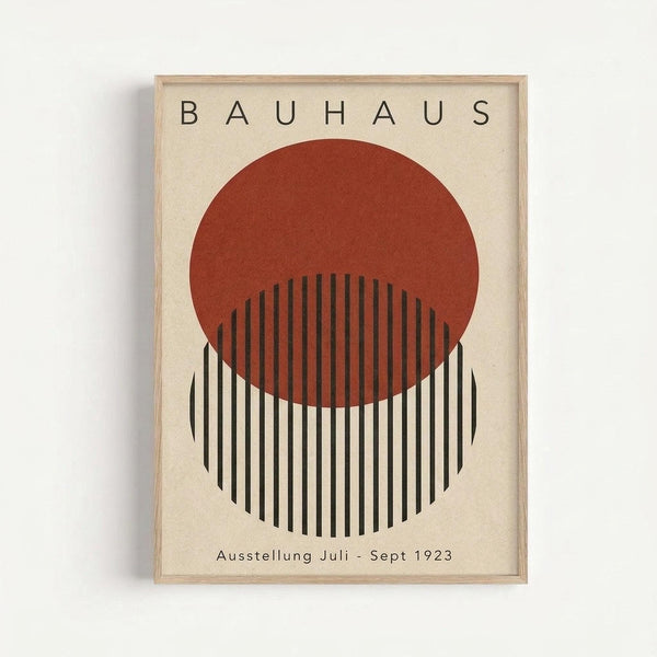 Minimalist geometric poster with cream background featuring a rust-colored circle overlapping a black and white striped semicircle. Text reads "BAUHAUS" at top and "Ausstellung Juli - Sept 1923" at bottom. The artwork is displayed in a light wooden frame against a white wall, showcasing clean Bauhaus design principles with bold geometric shapes and typography.