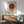 May include: Framed Bauhaus exhibition poster with rust red circle overlapping black and white striped circle on cream background, displayed above mid-century modern wooden credenza with white ceramic lamp, beside sage green upholstered armchair with wooden frame, natural lighting casting geometric shadows across white wall.