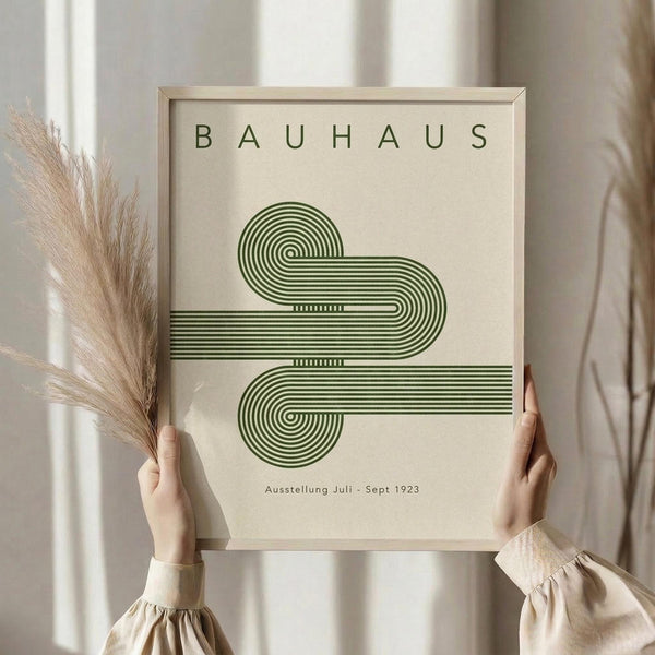 May include: Person holding framed Bauhaus poster with green geometric spiral design on cream background. Dried pampas grass visible to left. Neutral beige clothing and soft window lighting create minimalist styling. Wooden frame surrounds vintage-inspired exhibition print with German text.