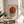 May include: Framed Bauhaus exhibition poster with geometric design featuring overlapping red and black striped circles on cream background, displayed on round wooden side table next to small potted plant, with pendant lights and beige chair visible in minimalist room setting