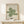 May include: Framed Bauhaus poster with green geometric spiral design made of curved parallel lines on cream background, displayed on light wood surface next to small wrapped white gift box with twine and greenery, leaning against white wall in minimalist setting