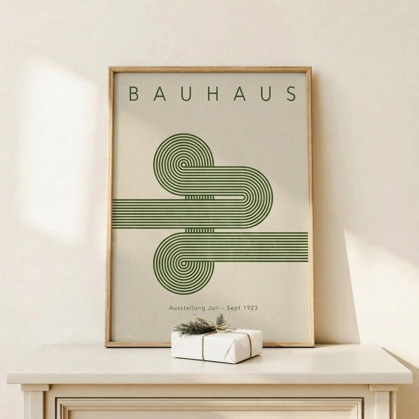 May include: Framed Bauhaus poster with green geometric spiral design made of curved parallel lines on cream background, displayed on light wood surface next to small wrapped white gift box with twine and greenery, leaning against white wall in minimalist setting