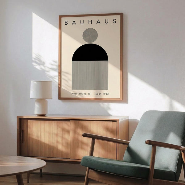 May include: Geometric Bauhaus-style poster with abstract shapes including black semicircle, striped rectangle, and concentric circle design, displayed in wooden frame above mid-century modern credenza with ceramic lamp, next to sage green upholstered armchair in bright, minimalist room with white walls and natural lighting casting plant shadows.