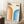 May include: Framed geometric poster with "BAUHAUS 1923" text and curved rainbow stripes in orange and blue gradients, displayed on white carpet against white paneled wall. Light wood frame, positioned next to wooden side table with white coffee cup and saucer. Beige knit throw blanket visible on right side. Natural lighting creates soft shadows.